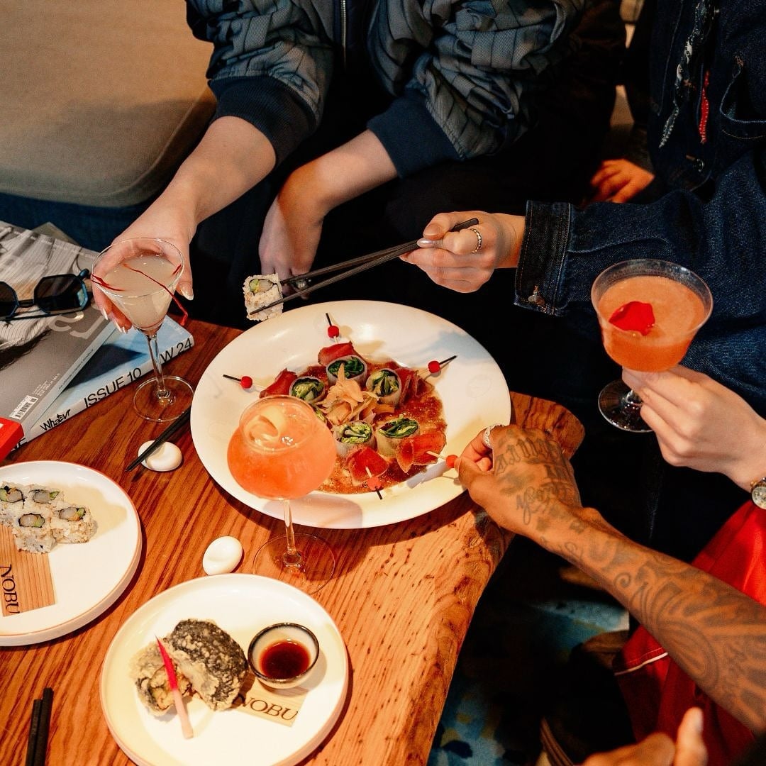 Group sharing sushi and cocktails at a wooden table, with colourful dishes and drinks."