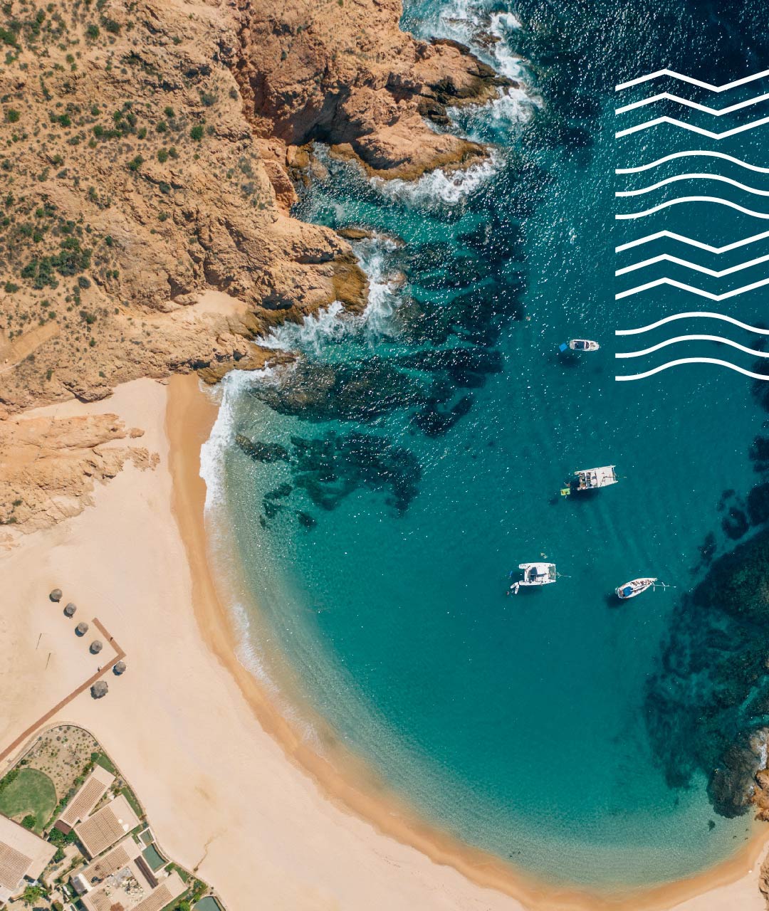 Aerial view of a sandy beach and rocky coastline with boats in clear blue water.