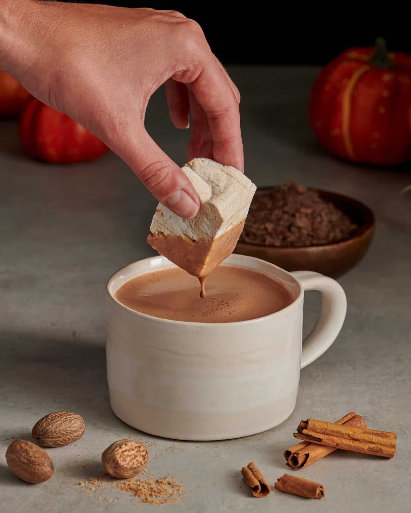 A hand dips a large marshmallow into a cup of hot chocolate, surrounded by cinnamon sticks and nutmeg.