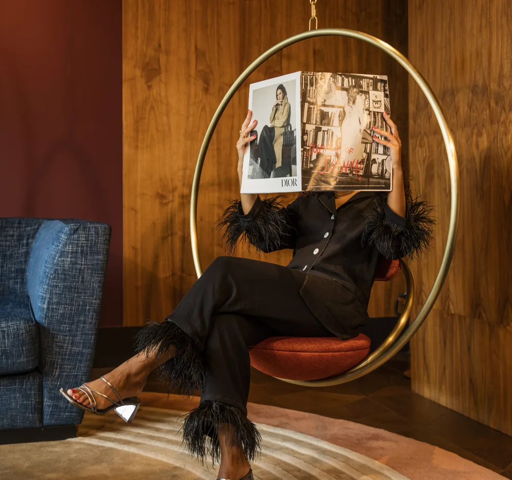 Person reading a Dior magazine on a circular swing chair, dressed in black loungewear with feather trim.